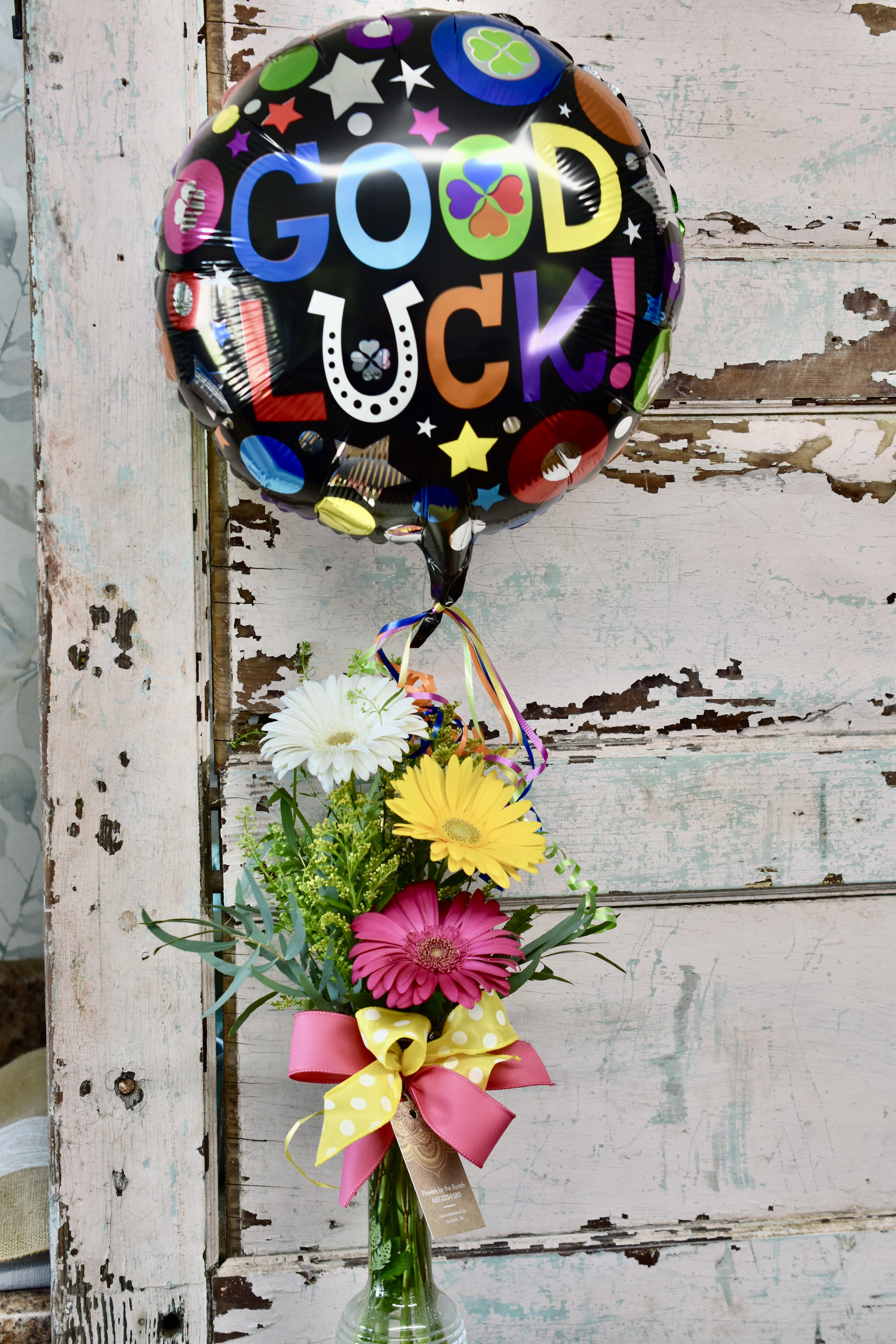 Tall vase of pink, yellow, and white gerbera daisies with a colorful round 'Good Luck' balloon.