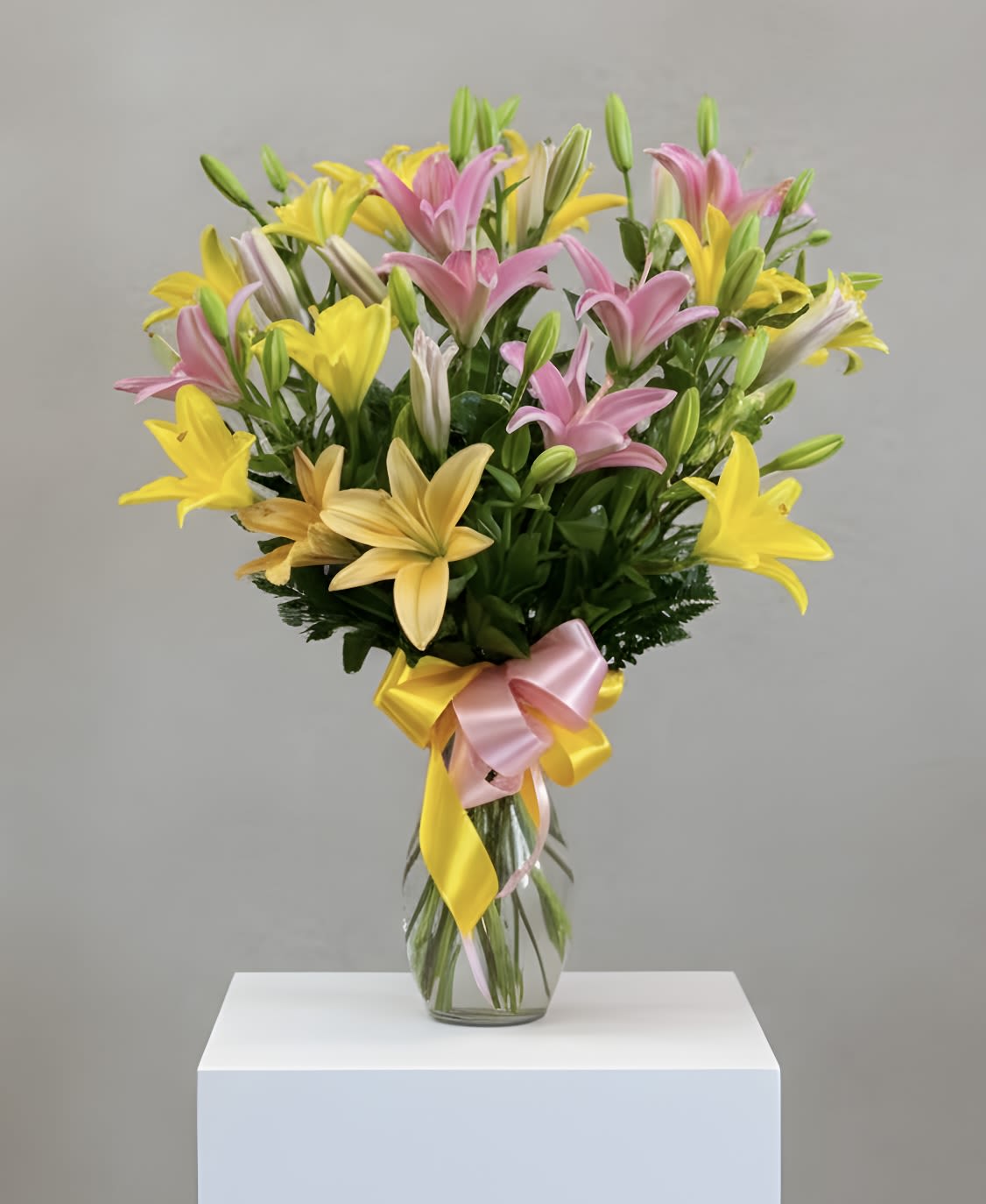 Tall clear vase of pink and yellow lilies with buds, tied with a pink and yellow ribbon bow.