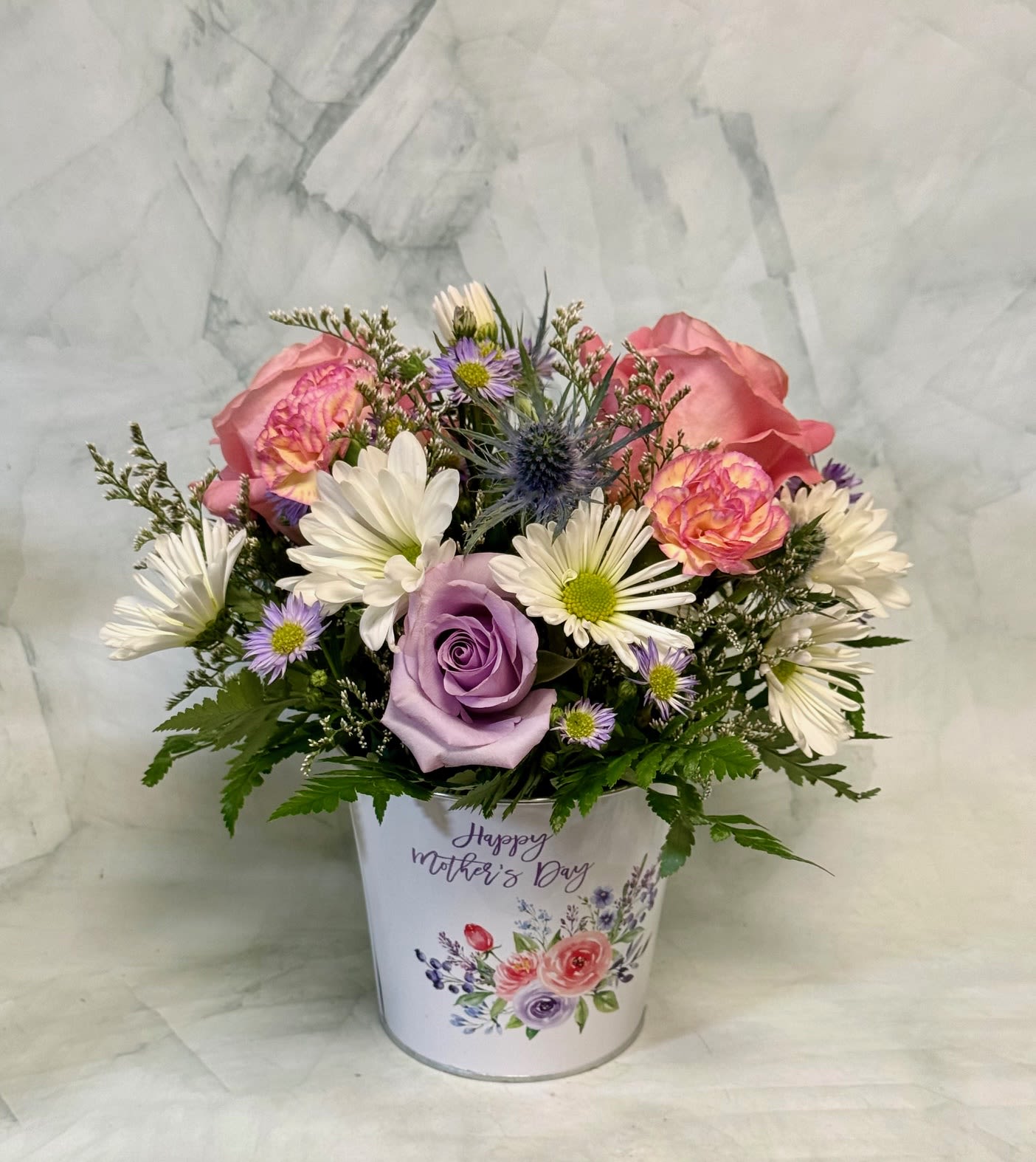 Happy Mother's Day Flower Pot - This arrangement is perfect to celebrate the mom in your life. The arrangement arrives in an adorable metal tin container that is in the shape of a flower pot. This container can be repurposed and serves as the perfect keepsake for Mother's Day. The arrangement includes roses, pom pons, mini carns, aster, thistle and limonium