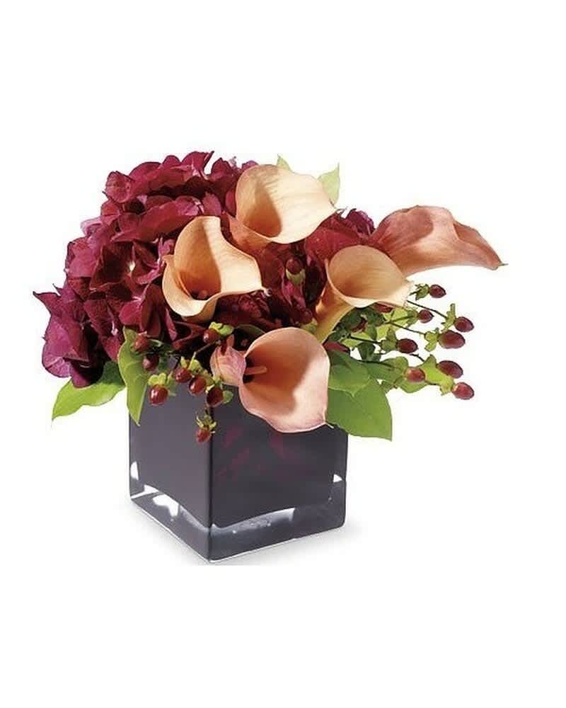 Low arrangement of peach calla lilies and burgundy flowers in a dark square glass vase