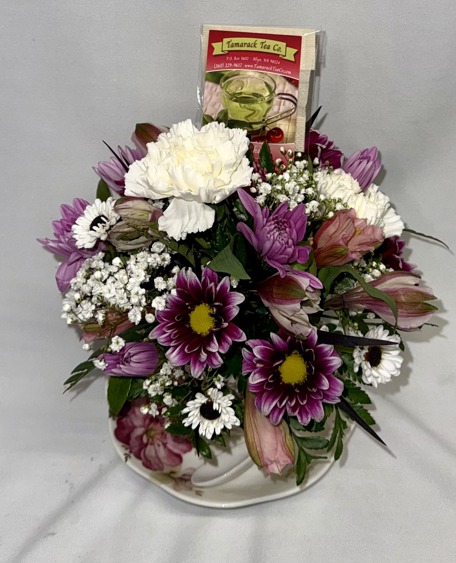 Tea party! - A cozy cluster of mixed roses, daisies, alstroemeria and chrysanthemums arranged in a low, rounded ceramic teacup and saucer makes a sweet birthday bouquet or Mother's Day gift that feels like a warm invitation to pause. This tea party flower arrangement with included Tamarack Tea Co. packets is also perfect for Get Well wishes, Thank You gestures, graduations, or a just-because surprise-the recipient will feel comforted and truly seen by the thoughtful pairing of fresh blooms and a cup of tea.  Flowers are arranged compactly to fill the floral-patterned teacup and matching saucer, with a ceramic spoon. The low profile makes it an easy bedside or desk accent; delicate filler blooms and unopened lily buds add soft texture while a few individually wrapped tea packets rise gently from the back to add color and a small consumable gift.  Choose to have us select an assortment of Tamarack Tea Co. flavors or specify your preference in the special instructions-available caffeinated options include Sir Earl Grey, Black Elderberry, and Chai. Decaf &amp; herbal choices include Starfruit Kiwi, Bourbon Street Vanilla, Chocolate Mint Rooibos, Angel Mist, Blue Mango, Amazing Apricot Honey, and Kyoto Cherry. This keepsake teacup arrangement arrives ready to brighten a spring or summer with warmth and charm.