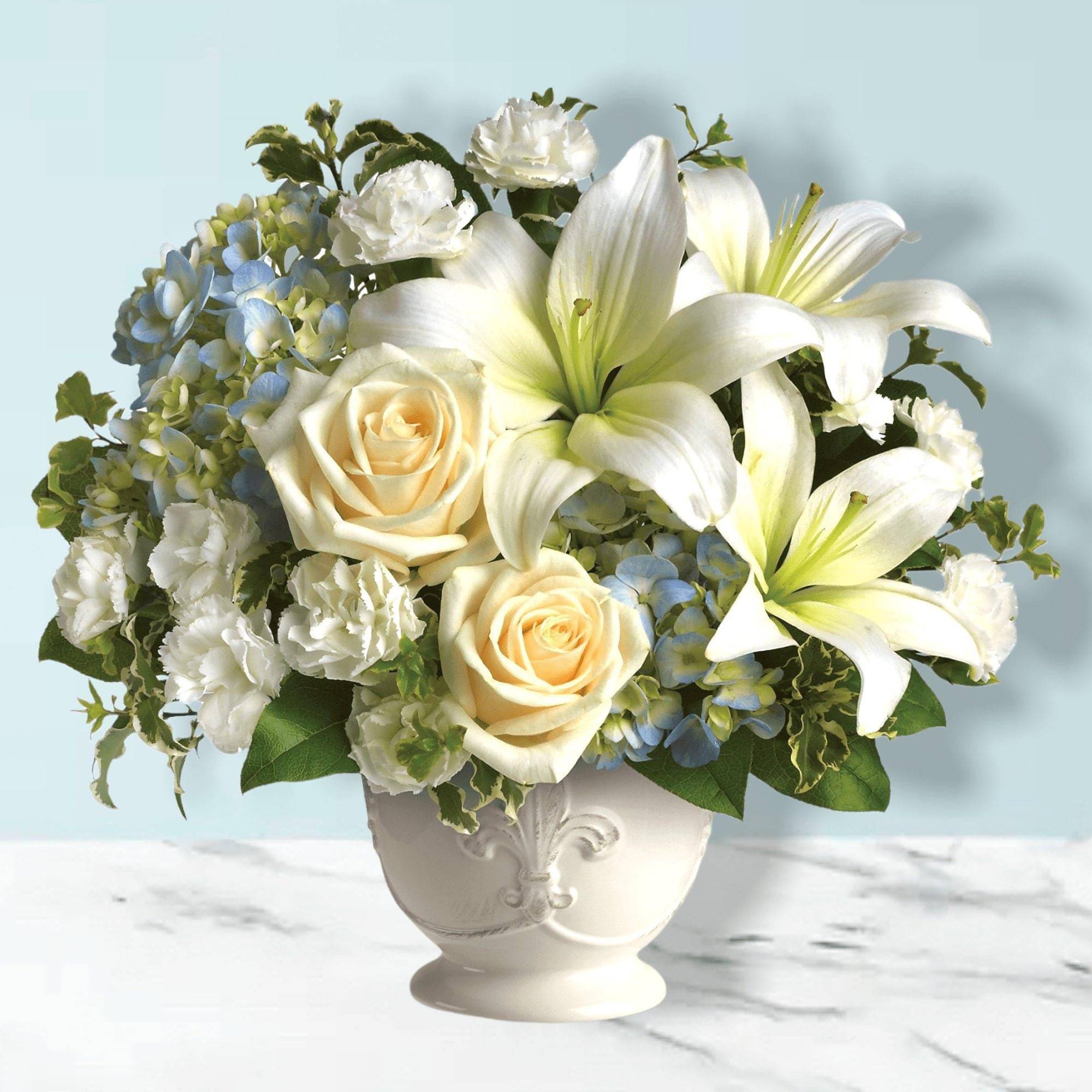White lilies and cream roses in a white vase with blue hydrangeas