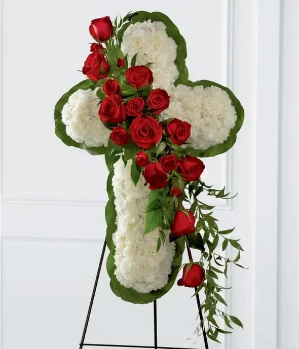 Standing floral cross of white blooms with cascading red roses on a metal easel.