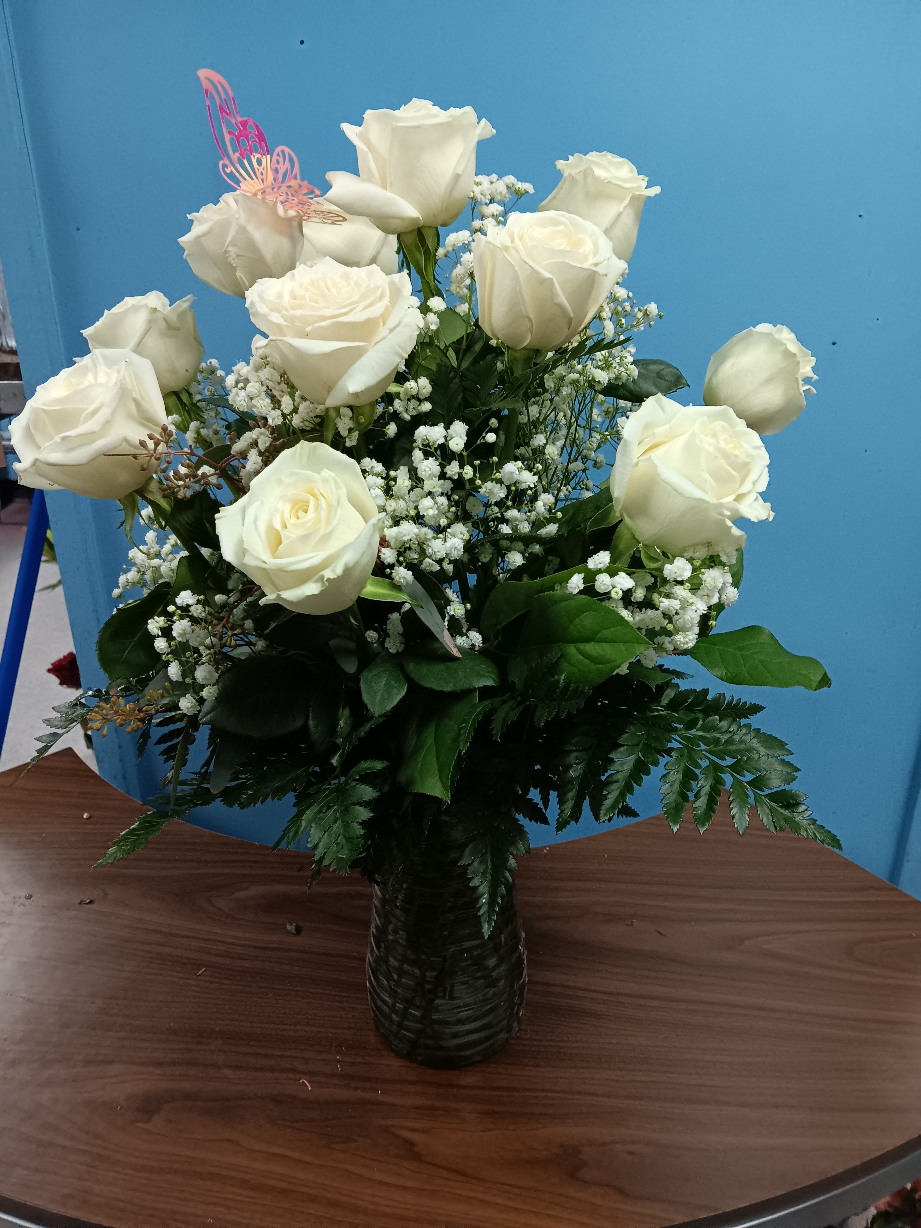 1 Dozen White Roses - Creamy white roses rise in a tall textured glass vase, their long stems gathered into a graceful, upright bouquet that feels both classic and softly romantic - perfect as a birthday bouquet, anniversary gift, or a sympathy arrangement for a memorial service. Sprays of delicate white filler blooms thread between the roses like little clouds, and a pastel decorative butterfly pick adds a gentle, whimsical touch that makes the person you're thinking of feel seen and comforted.  Lush dark-green foliage forms a full base that frames each bloom and heightens the creamy petals' glow. Available in three sizes - a simple dozen, Deluxe with 18 stems, and Premium with 24 stems - each comes arranged in the clear glass vase pictured. Ideal for Mother's Day, Valentine's Day, weddings, get-well wishes, or "just because" moments, these low-maintenance roses arrive ready to display; change the water every few days and trim stems for longer enjoyment.