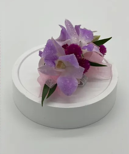 Lavender orchid corsage with bright purple accents and pink ribbon on a white round base.