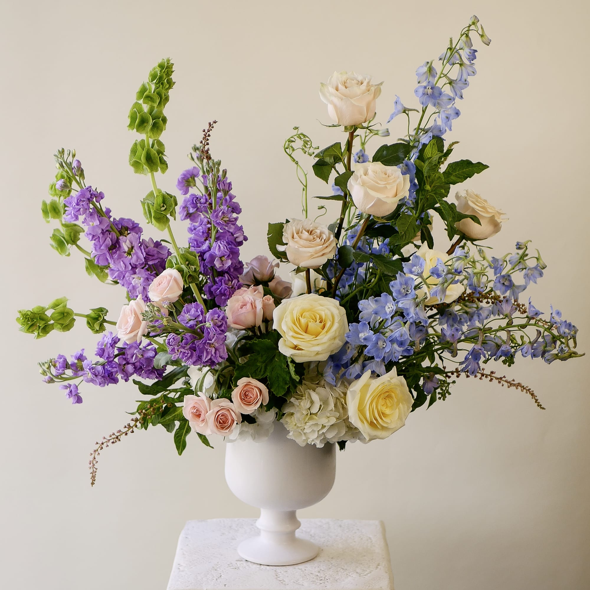 The English Garden - A striking, large-scale arrangement designed for moments that deserve presence. The English Garden layers purple stock, blue delphinium, and soft blush spray roses around a foundation of cream garden roses and white hydrangea — finished with sculptural green bells of Ireland that extend the design upward and outward.  Presented in a classic white pedestal compote, this arrangement commands attention without sacrificing softness. The cool violet and blue tones balance beautifully against warm cream and blush, creating a palette that feels both elevated and inviting. Ideal for milestone celebrations, corporate gifting, sympathy tributes, or as a statement centerpiece for the home.  The arrangement approximately measures 31" tall and 28" wide with flowers.