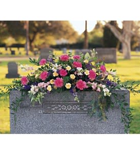 Graveside spray of pink, yellow, and white flowers draping over a gray headstone.