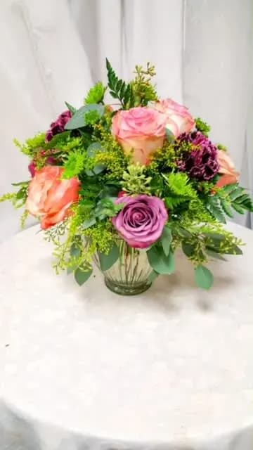 Round arrangement of peach and lavender roses with dark plum flowers in a clear glass vase