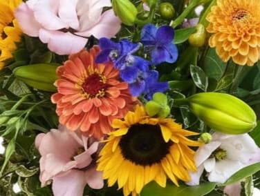 Bright mixed bouquet with sunflowers, orange blooms, pink lisianthus, blue delphinium, and lily buds.