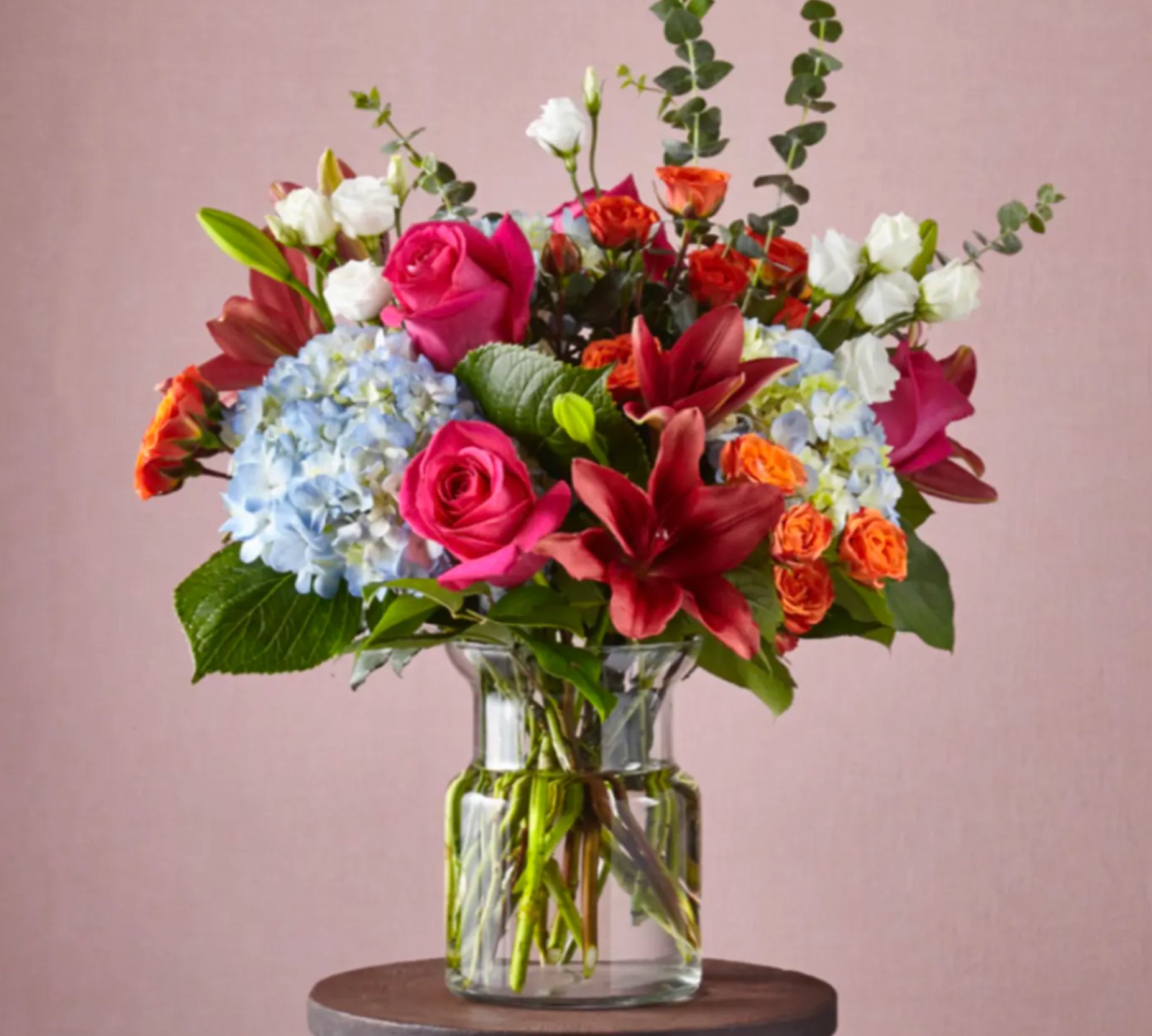 Mixed bouquet with pink roses, red lilies, blue hydrangeas and orange spray roses in a clear glass vase