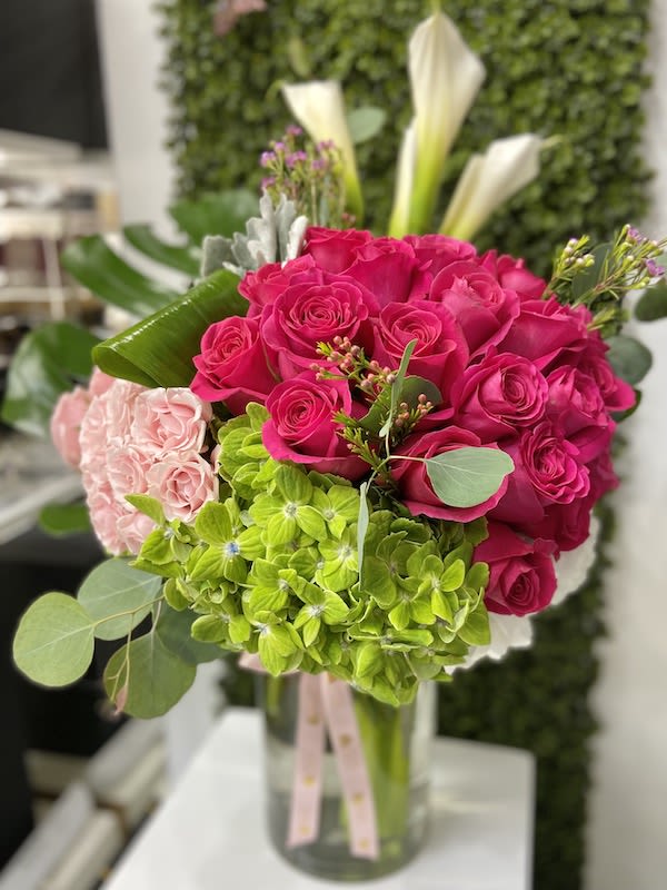 Bright pink roses, green hydrangeas, and pale pink blooms arranged in a clear glass vase with ribbon
