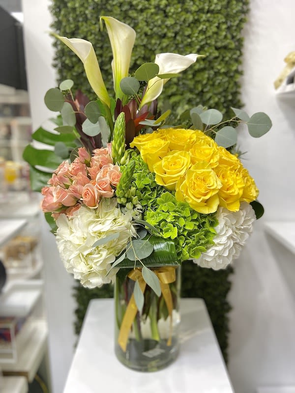 Tall arrangement of yellow roses, peach spray roses, white hydrangeas, and calla lilies in a glass vase