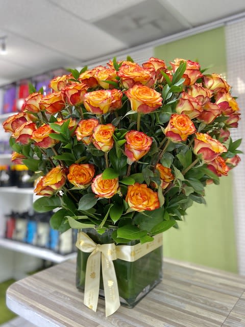 Large arrangement of orange and yellow roses in a square glass vase with a cream ribbon bow.