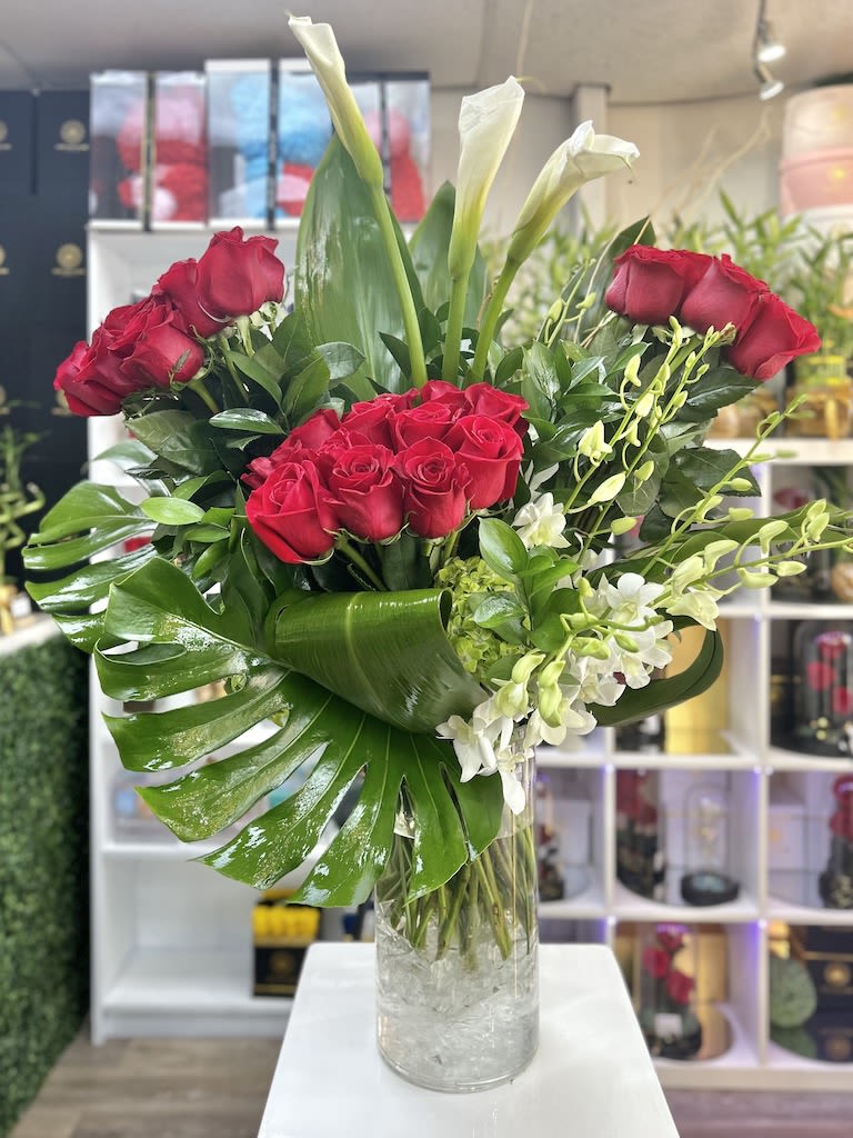 Tall arrangement of red roses, white calla lilies, and orchids in a clear glass vase with large glossy leaves