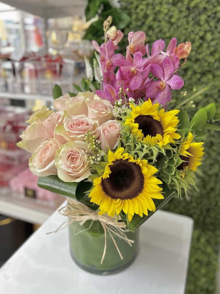 Arrangement of pale pink roses, yellow sunflowers, and pink orchids in a glass vase tied with raffia.