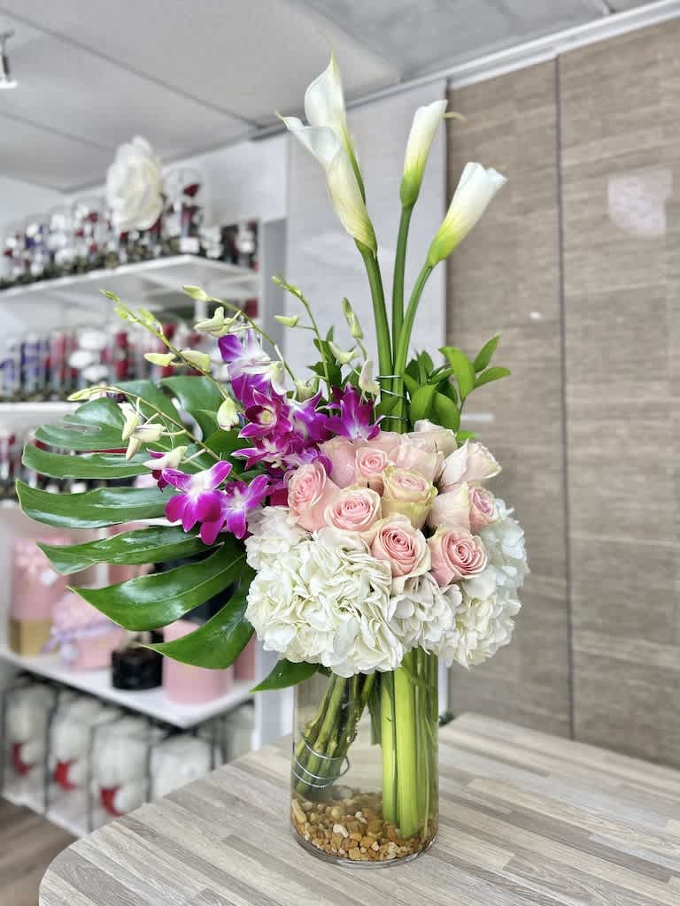 Tall mixed arrangement with white hydrangeas, pink roses, purple orchids and calla lilies in a clear glass vase