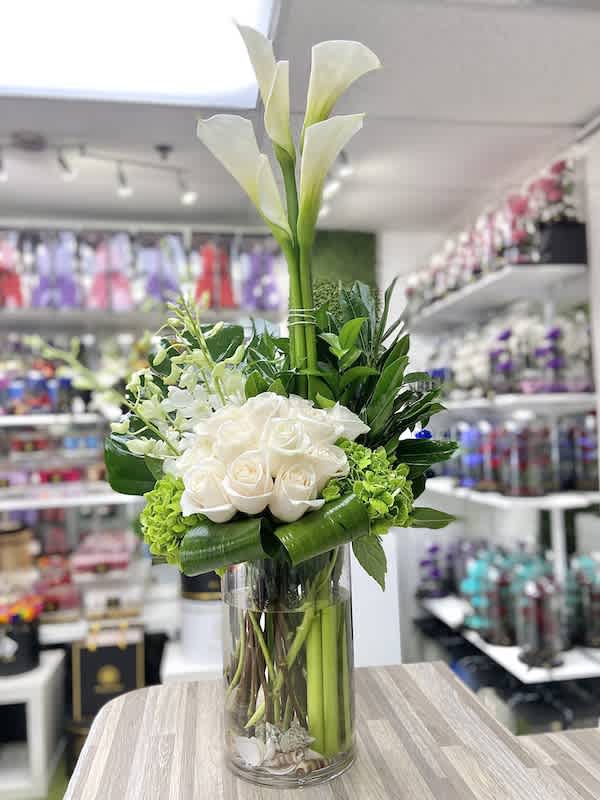 Tall white calla lilies and white roses with green accents arranged in a clear glass cylinder vase.