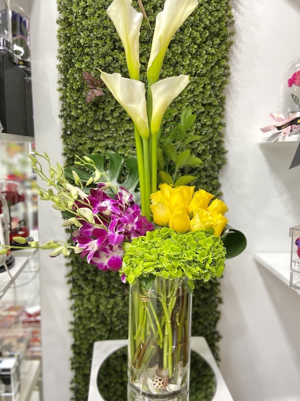 Tall arrangement of white calla lilies, green hydrangea, yellow roses, and purple orchids in a clear glass vase