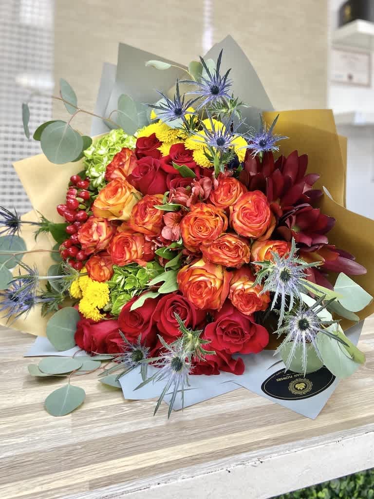 Hand-tied bouquet of red and orange roses with yellow blooms and blue thistles in kraft paper wrap