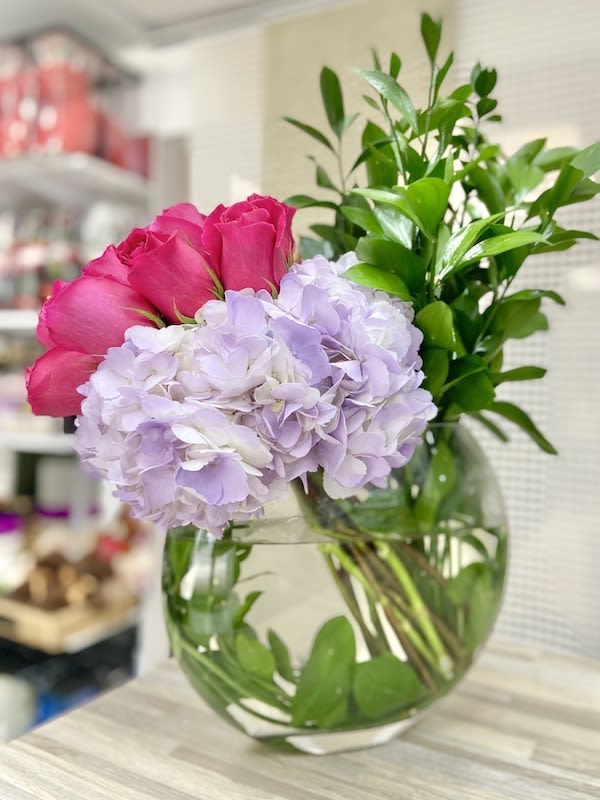Round glass vase with pink roses, pale lavender hydrangeas, and tall green foliage.