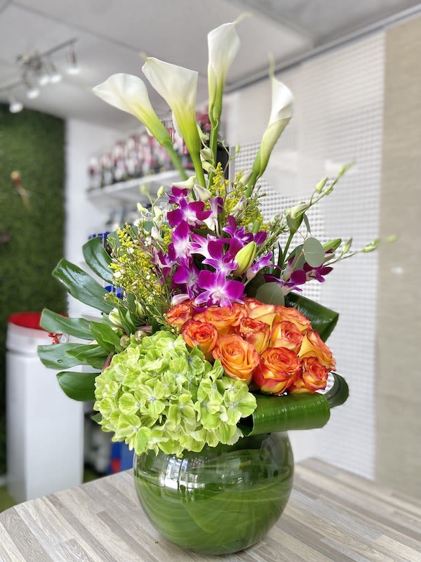 Tall mixed arrangement with calla lilies, orchids, roses, and hydrangea in a leaf-lined glass vase.