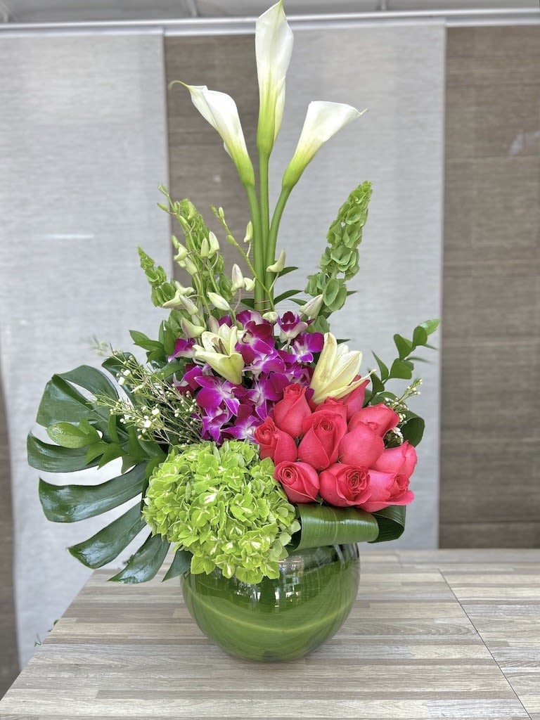 Tall arrangement of white calla lilies, pink roses, orchids, and green hydrangea in a round glass vase