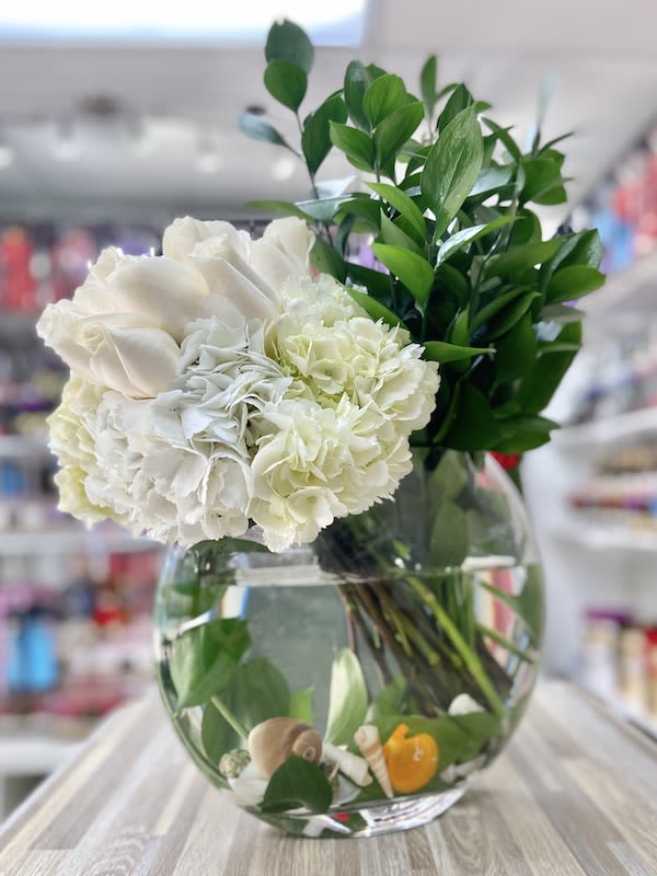 Round glass vase with white roses and hydrangeas, tall greenery, and seashells with a small yellow duck in the water