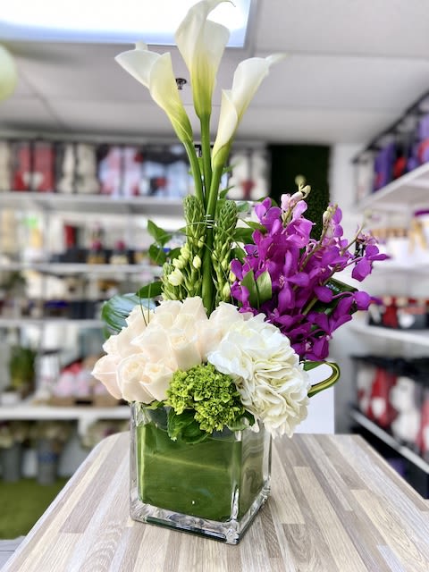 Tall white calla lilies with purple orchids and pale roses in a green-lined glass cube vase.
