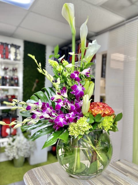 Tall mixed arrangement with white calla lilies, purple orchids, and orange blooms in a round glass vase