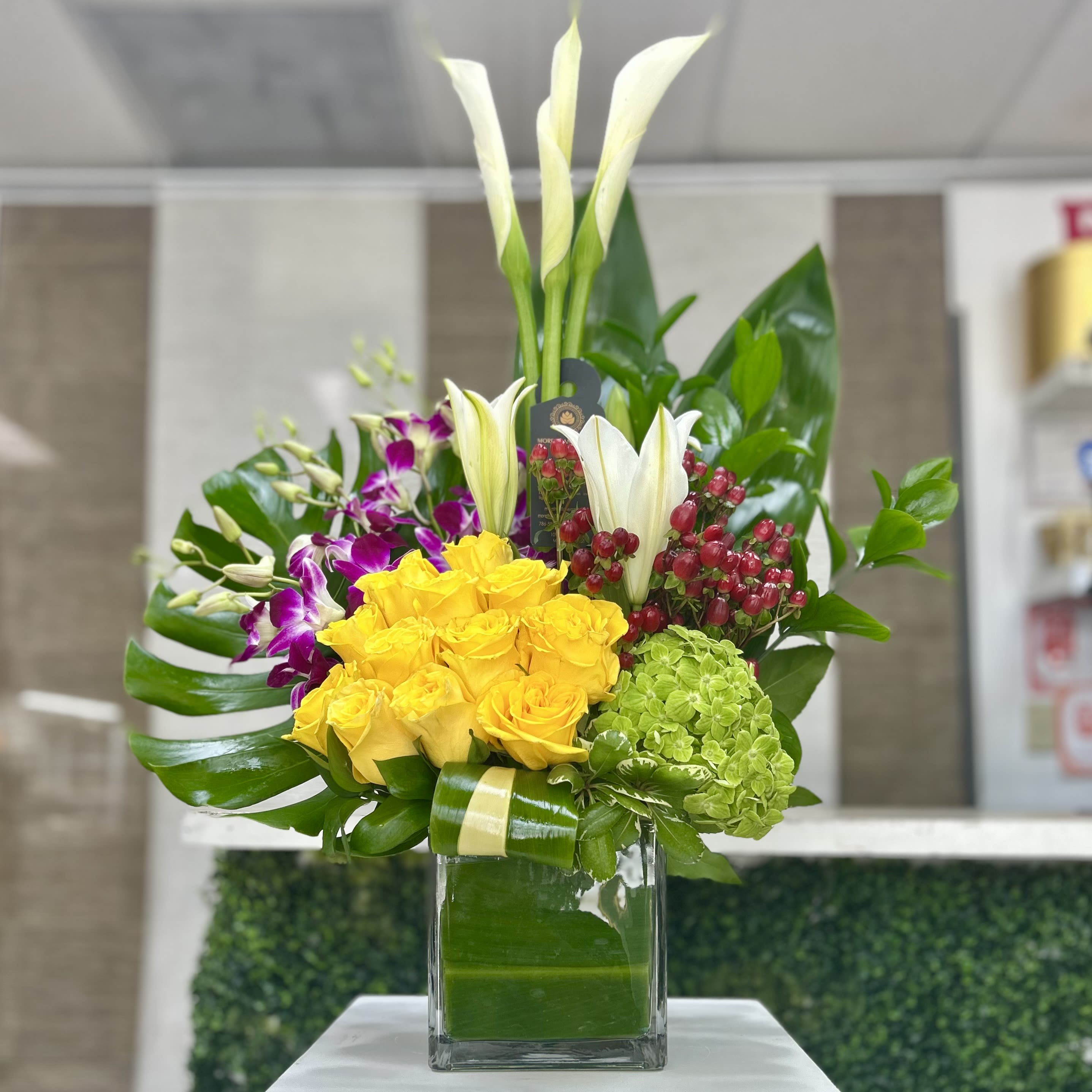 Tall modern arrangement with yellow roses, white lilies, purple orchids, and green hydrangea in a glass cube vase