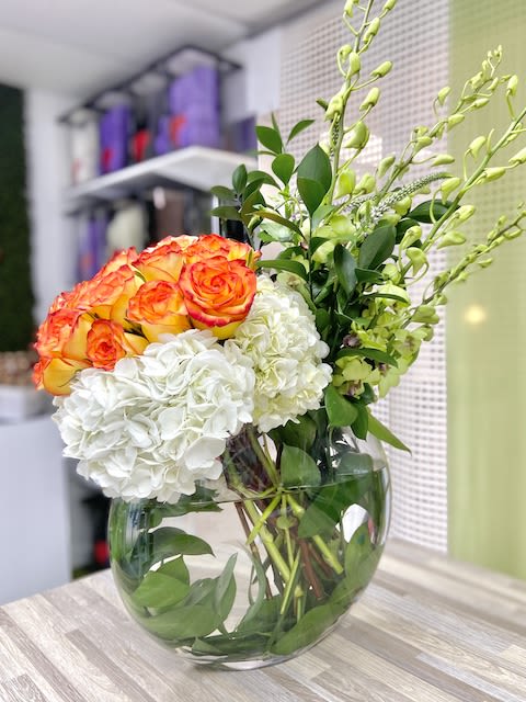 Round glass vase with orange roses, white hydrangeas, and tall green orchid stems