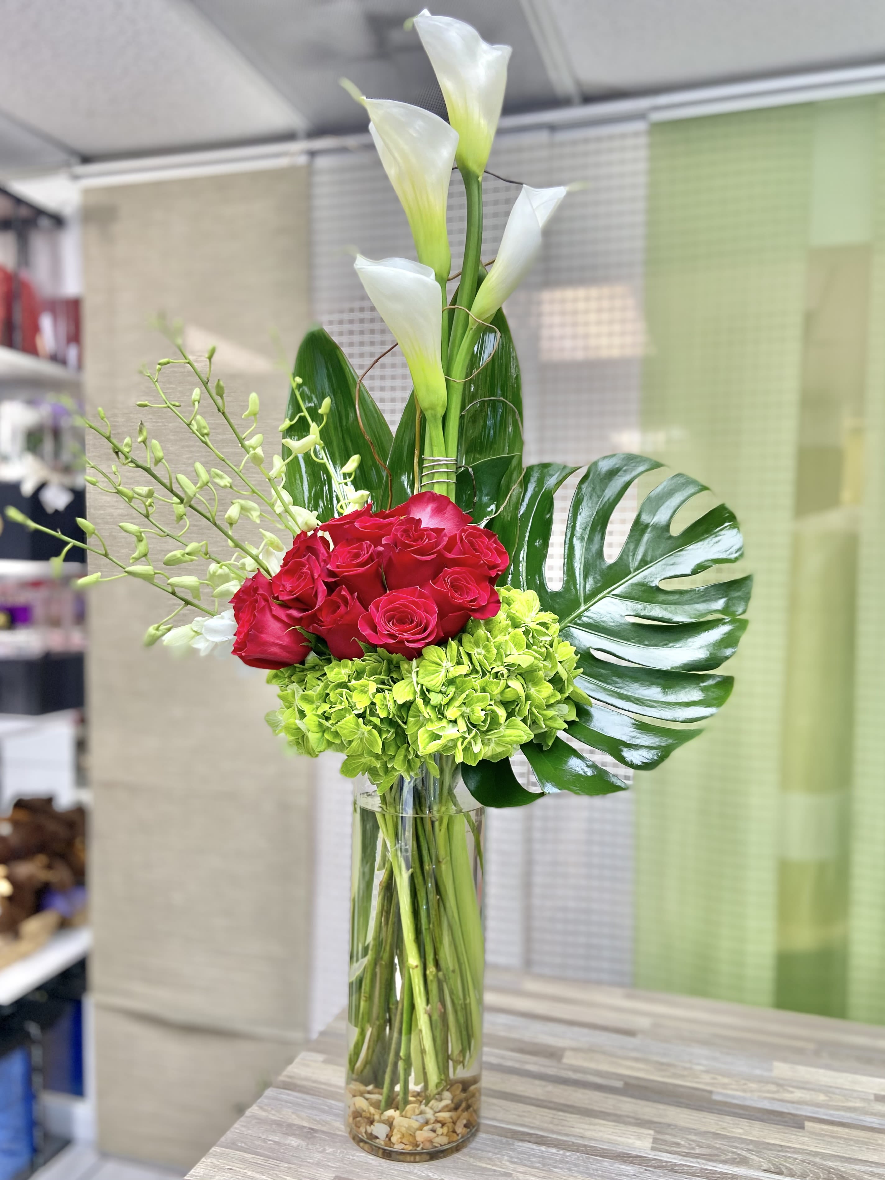 Tall arrangement of white calla lilies, red roses, and green hydrangeas in a clear glass cylinder vase