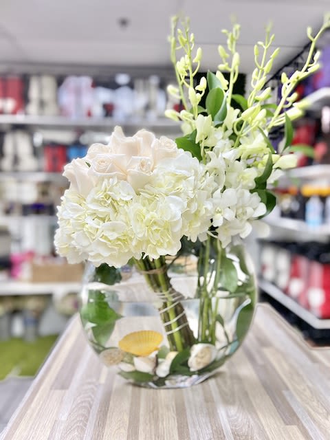 Round glass vase of white roses, hydrangeas, and orchids with seashells in the water