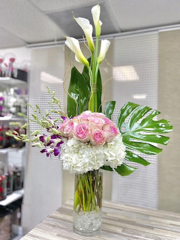 Tall arrangement of white calla lilies, pink roses, orchids, and hydrangeas in a clear cylinder vase.