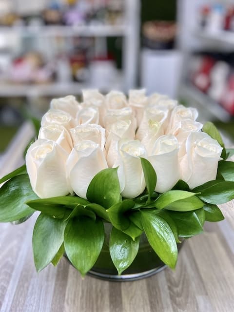 Low arrangement of cream roses tightly packed in a round glass vase with a collar of glossy green leaves.