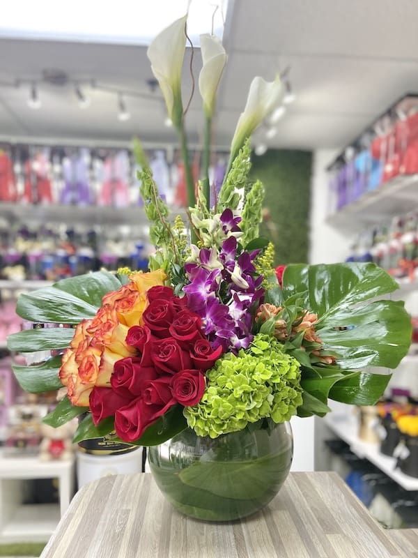 Tall arrangement of red roses, calla lilies, orchids and green hydrangea in a round glass vase with large tropical leaves.