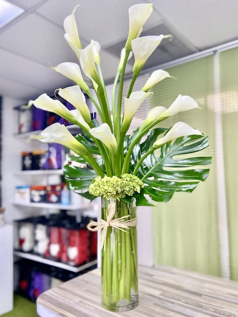 Tall arrangement of white calla lilies with large glossy leaves in a clear glass vase.