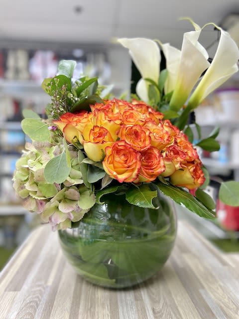 Low round arrangement of orange roses, green hydrangea, and white calla lilies in a glass bowl vase