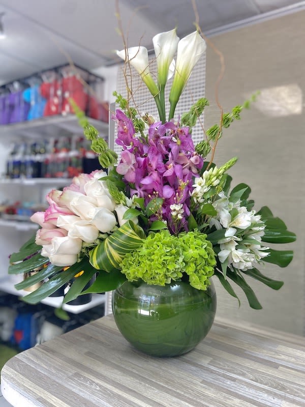 Tall arrangement of white calla lilies, roses, orchids and green hydrangea in a round green glass vase with bold leaves