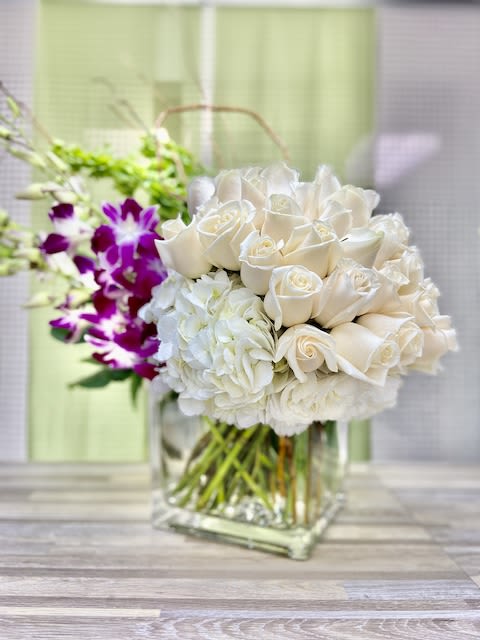 White roses and hydrangeas in a clear cube vase with purple orchids and green Bells of Ireland in the background.