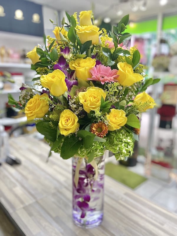 Tall arrangement of yellow roses, pink daisies, and orchids in a clear cylinder vase