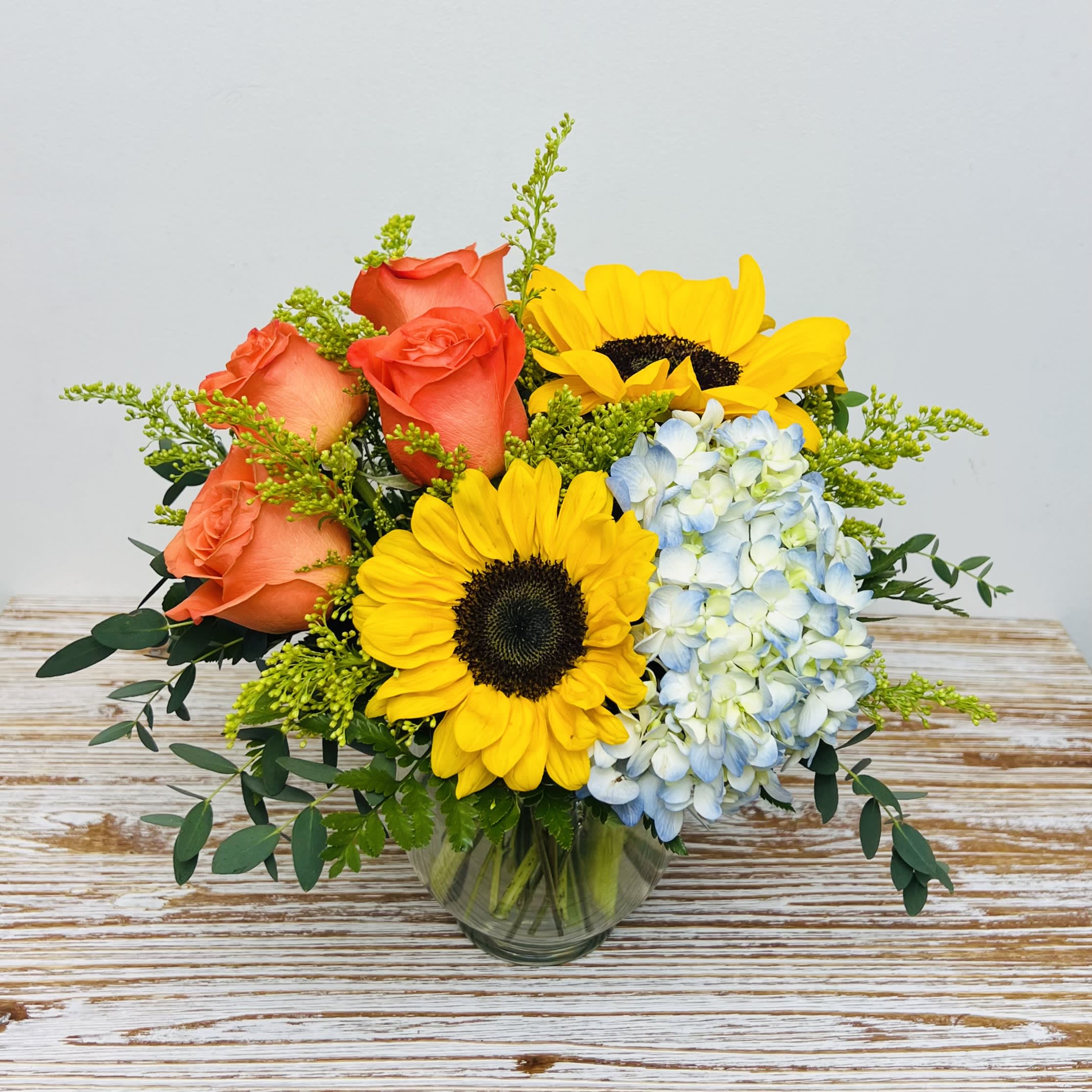 Daydream  - An arrangement that makes you want to daydream when gazing at it. Bold orange Roses, bright and lively Sunflowers, and sky blue Hydrangea are delightfully arranged with accents in a clear bowl.   Approximately 10” tall x 10” wide  Container Country of Origin: USA Flower Country of Origin: Ecuador and/or Columbia