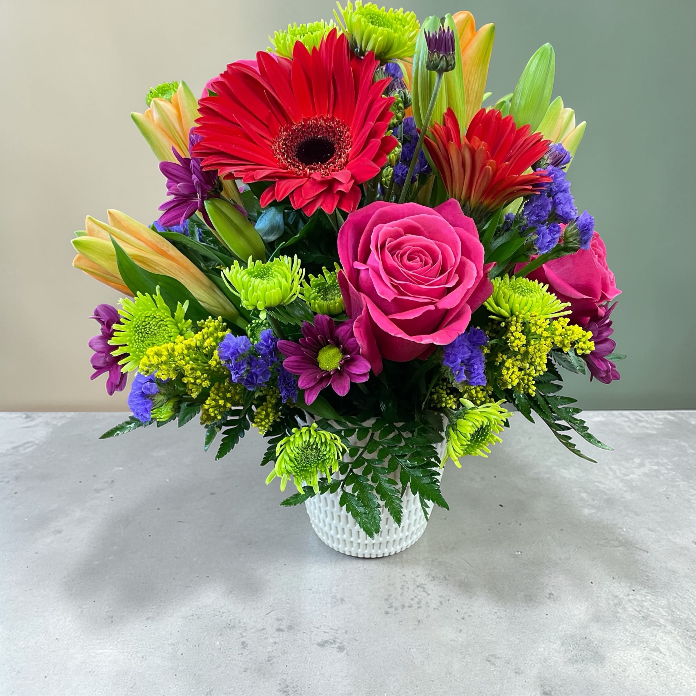Color, Color Everywhere  - A jubilant burst of hot pink roses and red gerbera daisies makes this flower arrangement an instant mood-lifter-perfect as a birthday bouquet, a Mother's Day surprise, or a bright "just because" gift to celebrate someone special. This mixed-color flower arrangement channels spring and Easter cheer without feeling seasonal-only, so it's equally right for graduations, get-well wishes, anniversaries, or simply letting someone know you're thinking of them.  Tightly gathered in a clean white textured metal vase, the design pairs  asiatic lilies with purple daisies, bright green button mums, and clusters of yellow filler blooms for lively contrast and depth. Small purple accent flowers weave through the rounded silhouette, creating a playful mix of shapes and saturated hues that reads joyful from every angle. The compact size fits tabletops and desks easily and arrives ready to display-no fussy assembly required-making it a thoughtful gift for housewarmings, thank-yous, new babies, or to brighten a hospital room.
