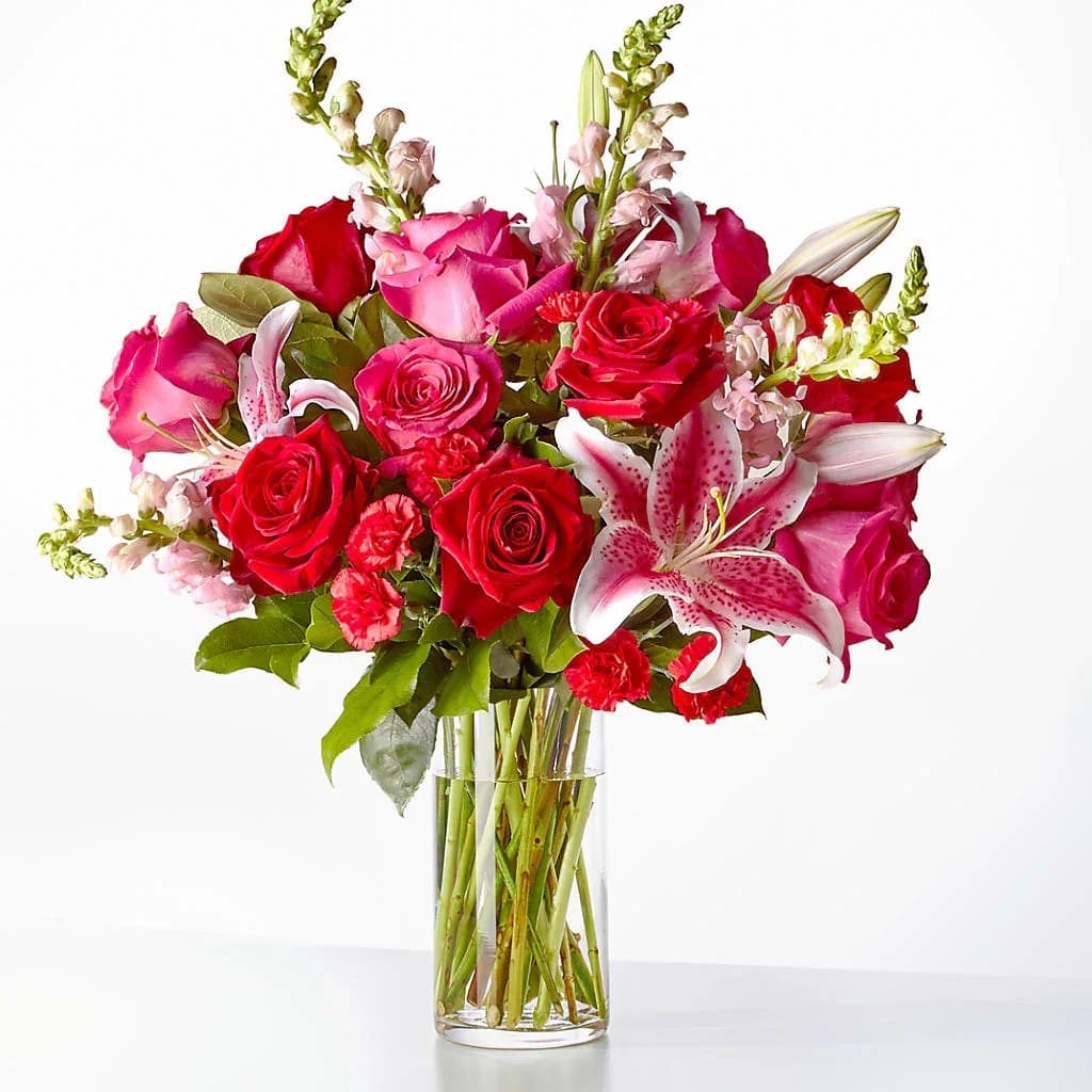 Arrangement of red and pink roses, lilies, carnations, and snapdragons in a clear glass vase