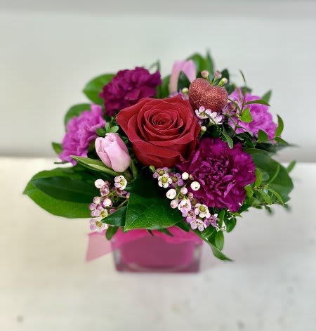 Compact arrangement of red rose, purple carnations and pink blooms in a pink cube vase with a glitter heart pick