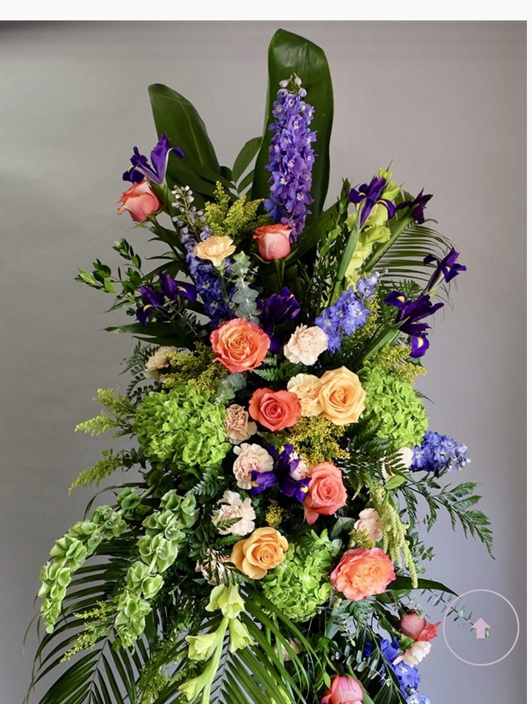 Tall standing arrangement of orange roses, purple delphinium, irises, and green hydrangeas with bold tropical foliage.