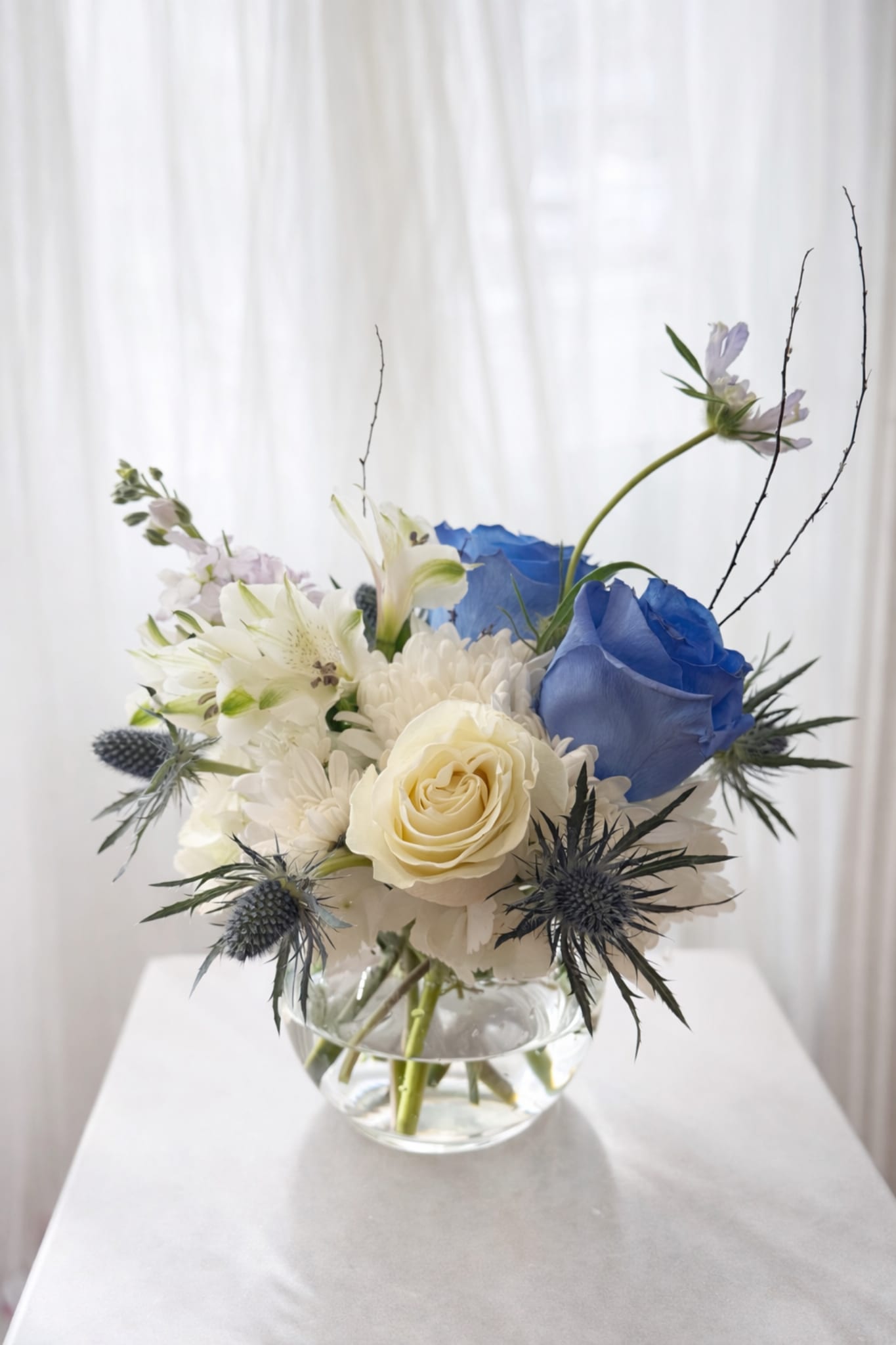 Azure Serenity Bowl - A delicate, modern floral arrangement presented in a clear glass bowl, showcasing a soft and airy design. The composition features elegant white blooms—including roses, chrysanthemums, and lilies—paired with a striking blue rose that adds a unique and contemporary touch. Accents of blue thistle bring texture and contrast, while light, wispy stems and seasonal fillers create a sense of movement and natural flow.  Set against a clean, softly lit background, the arrangement feels fresh, refined, and calming—perfect for a minimalist aesthetic or a thoughtful, sophisticated gift.