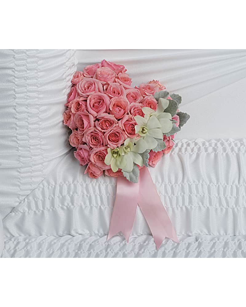 Heart-shaped pink rose arrangement with white orchids and pink ribbon on white ruched fabric.