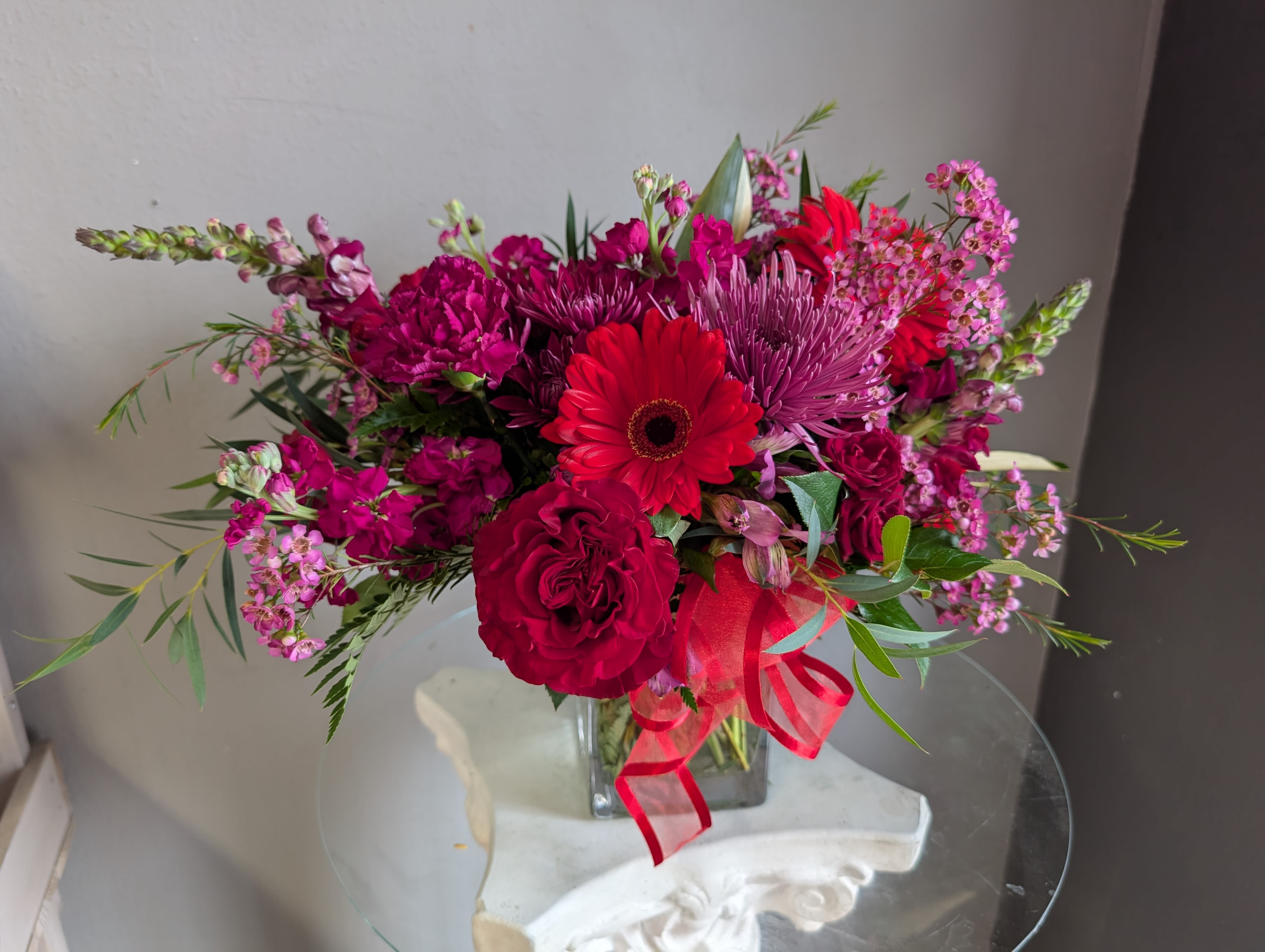 Vibrant arrangement of red and magenta blooms in a clear glass cube vase with a red ribbon bow.