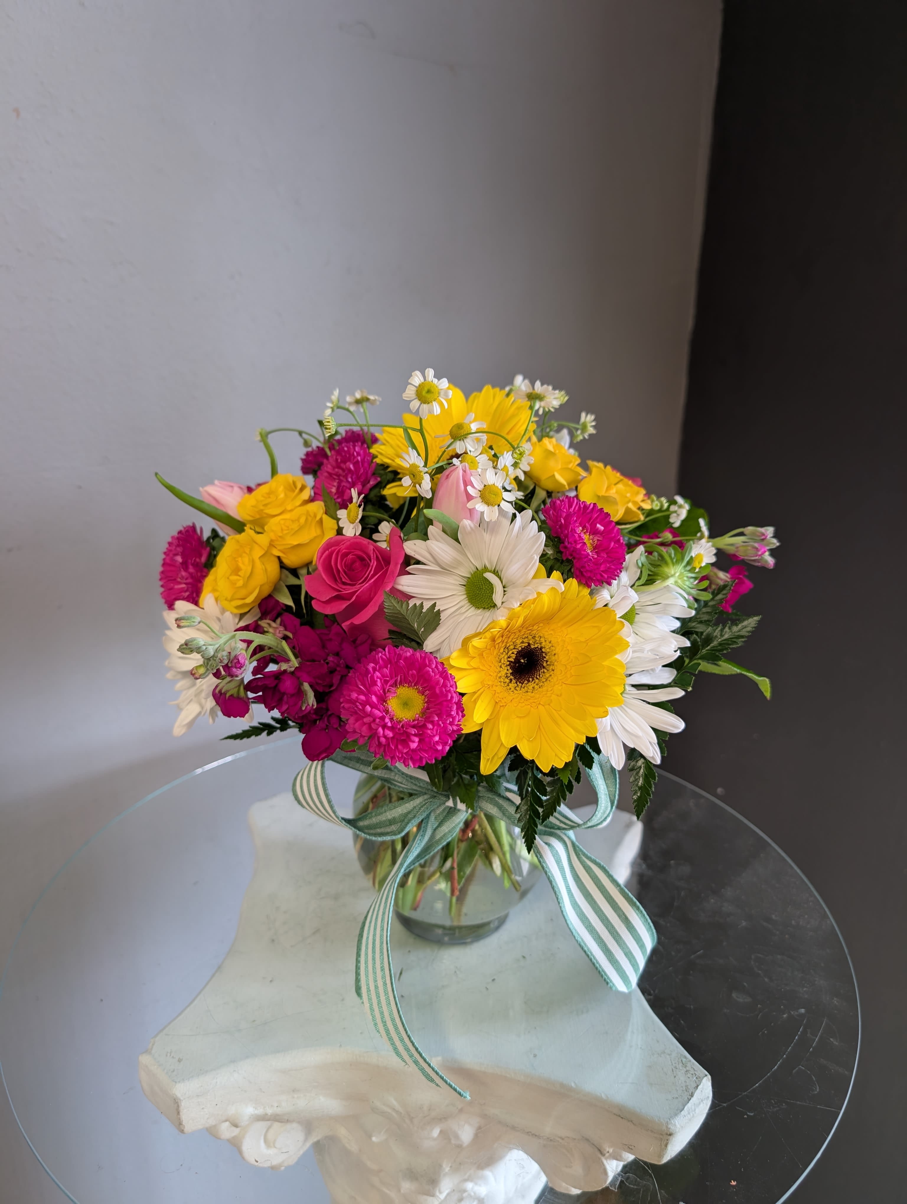 Colorful bouquet of yellow gerbera, pink and white daisies, and roses in a clear vase with striped ribbon.